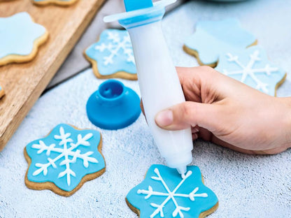 stylo déco blanc et bleu rempli de glaçage blanc tenu dans la main d'une femme et utilisé pour décorer des biscuits de Noël avec du glaçage bleu et blanc en forme de flocon de neige.