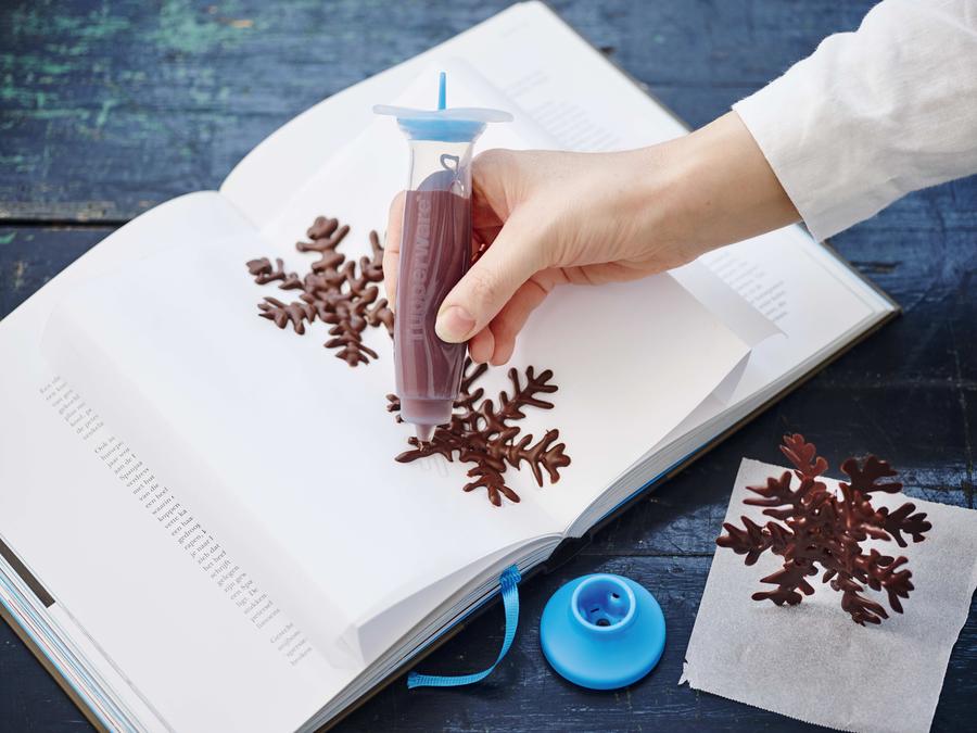 femme tenant le stylo deco tupperware avec du glaçage au chocolat et l'utilisant pour faire des desserts de flocons de neige en chocolat sur une serviette de table placée dans un livre